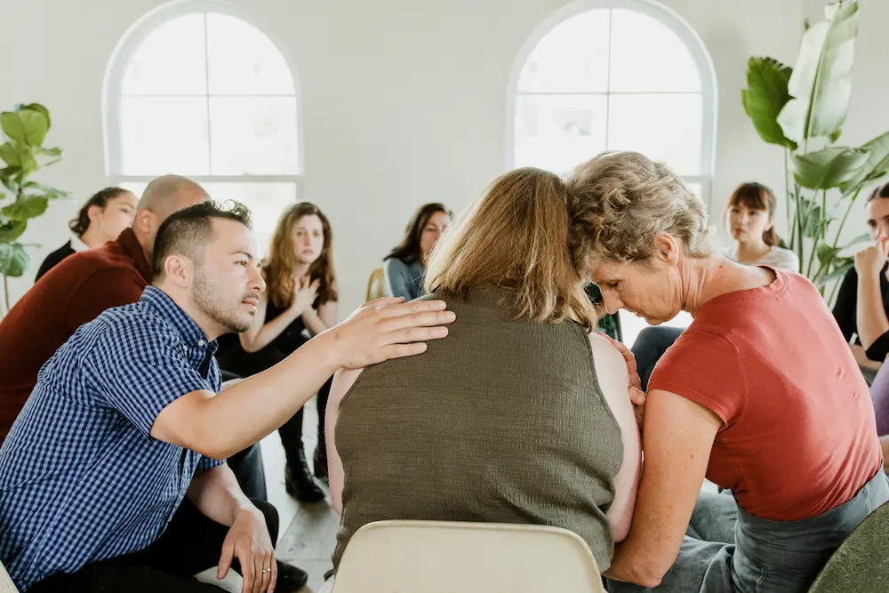 A diverse group of adults sits in a circle during a support group session in a brightly lit room with arched windows. In the foreground, a man in a blue checkered shirt leans forward to place a comforting hand on the back of a woman whose back is to the camera. To her right, an older woman in a red shirt leans her head gently against the center woman's head and shoulder. In the background, other group members watch the interaction with empathetic and serious expressions.